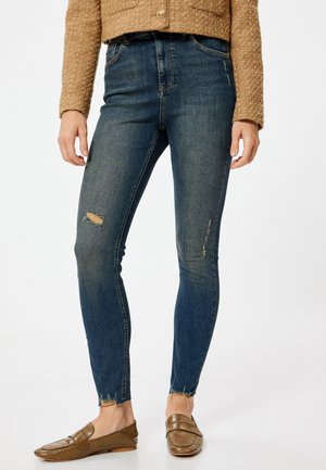 Jeans skinny de un azul oscuro con detalles desgastados, que presentan un dobladillo deshilachado y bolsillos, combinados con unos mocasines marrones texturizados.