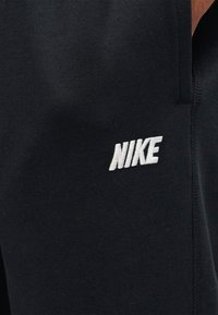 Μαύρο ύφασμα με λευκό κεντητό λογότυπο Nike στην περιοχή του άνω μηρού ενός ενδύματος, που δείχνει ραμμένες λεπτομέρειες της ραφής.