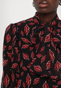 Blouse noire transparente avec motif de feuilles rouges et blanches, manches bouffantes et nœud noué au cou, portée par une personne à la peau foncée.