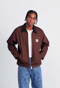 Brown jacket with a black collar, front zipper, and logo patch. Worn over a white shirt with light blue patterned jeans.