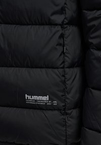 Svart puffjacka med kviltad textur, med varumärket "hummel" i vitt nära fållen och en dragkedja med accent. Slät tygfinish.