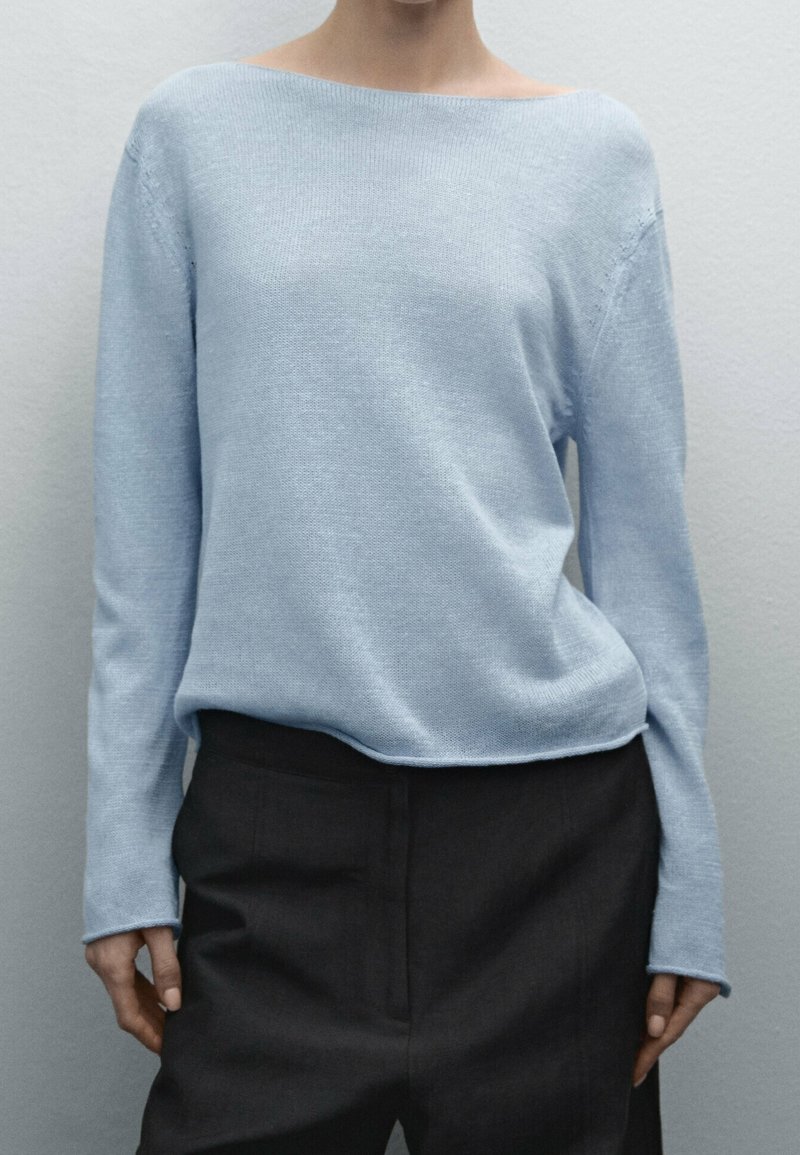 Light blue knit sweater with a wide neckline and long sleeves, paired with dark trousers. Smooth texture and relaxed fit.