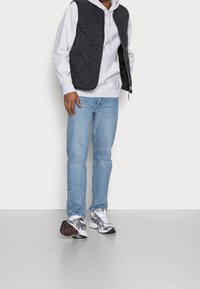 Černá prošívaná vesta přes šedou mikinu s kapucí, kombinovaná se světle modrými džínami a kovově stříbrnými teniskami s černými doplňky.