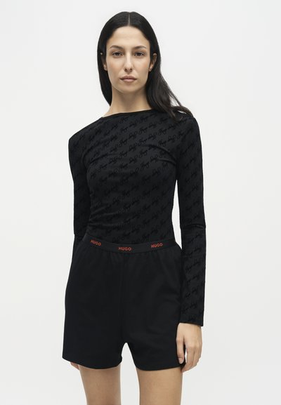 Femme aux longs cheveux foncés portant un haut à manches longues noir à motifs et un short noir avec une ceinture rouge avec le logo "HUGO".
