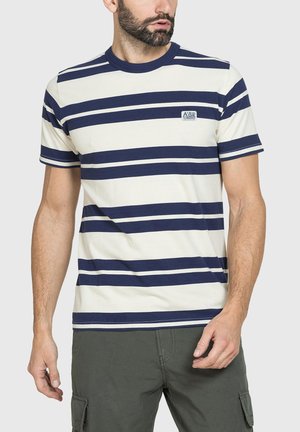 Uomo che indossa una maglietta a maniche corte a righe crema e blu navy con un piccolo logo sul petto, abbinata a pantaloncini cargo verde oliva.