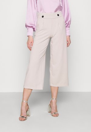 Lichtbeige wijde broek met hoge taille, voorzien van twee zwarte knopen en een zachte, lichte stof. Gecombineerd met zilveren hakken.