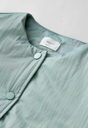 Țesătură texturată de culoare albastru deschis, cu un luciu neted. Are un decolteu rotund și două nasturi asortați. Eticheta din interior spune "NEXT."