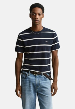 Junger Mann, der ein kurzärmeliges gestreiftes Hemd in Marineblau und Weiß, hellblaue Jeans und einen dunkel geflochtenen Gürtel trägt.