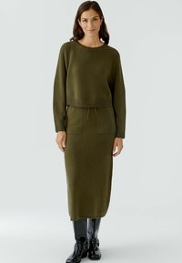 Olijfgroene gebreide twee-delige outfit, bestaande uit een verkorte trui met een ribbelzoom en een bijpassende midi-rok met twee voorkruiden.
