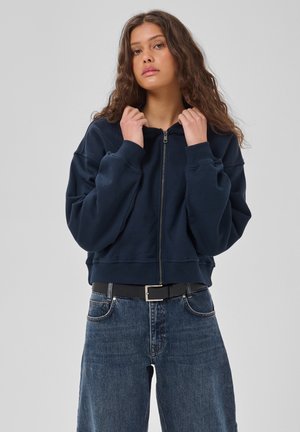 Jeune femme aux longs cheveux bouclés portant un sweat à capuche zippé bleu marine et un jean bleu, tenant le col du sweat à capuche avec les deux mains.