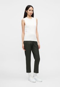 Γυναίκα με λευκό τοπ χωρίς μανίκια, σκούροι cropped παντελόνια, λευκές κάλτσες και λευκά αθλητικά παπούτσια, στέκεται μπροστά σε απλό φωτεινό φόντο.