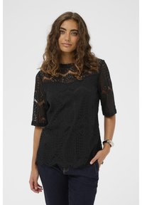 Fekete csipke felső, amelynek fodros szegélye, rövid ujjai és részletes virágmintái vannak. Sötét farmernadrággal és ezüst karkötővel párosítva.