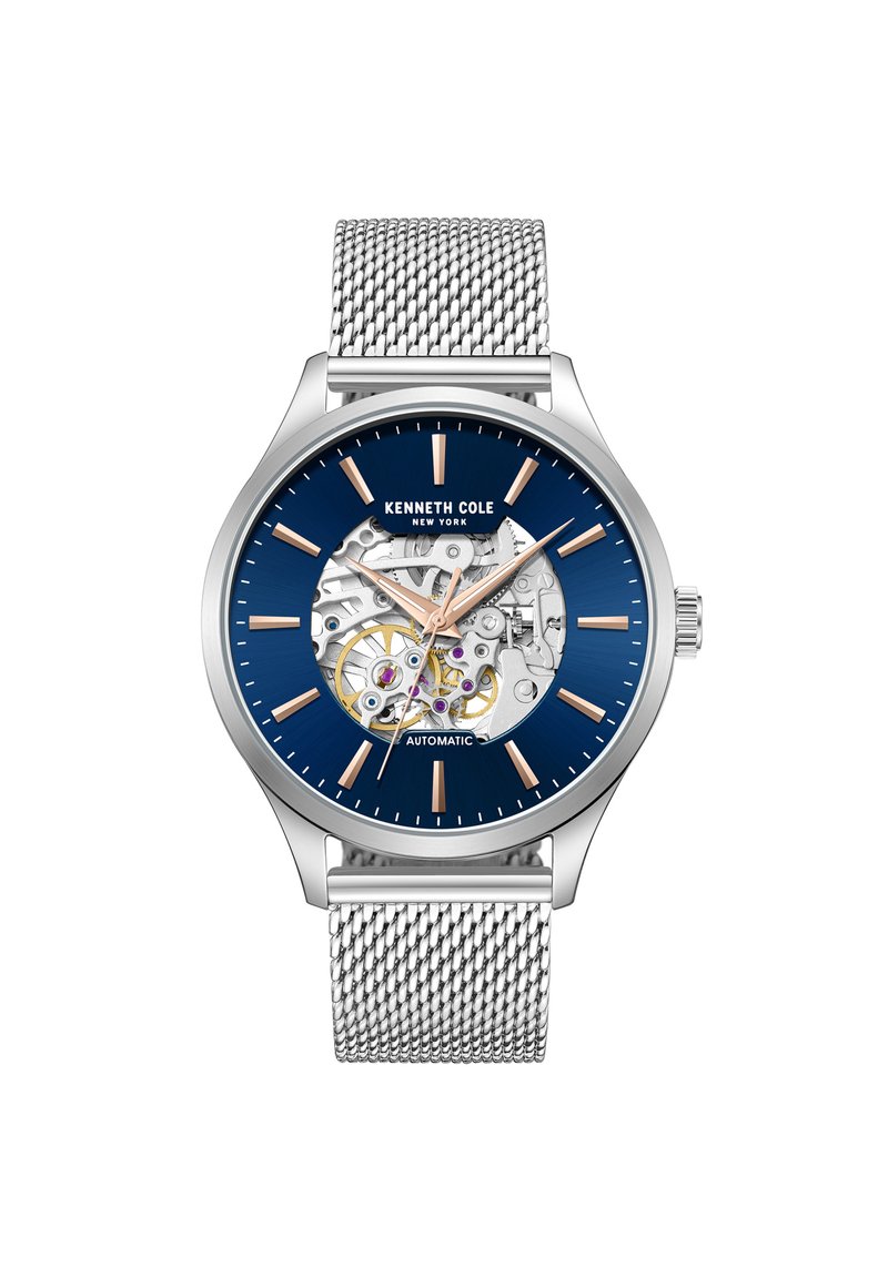 Reloj automático con caja de acero inoxidable, esfera azul, correa de malla plateada y mecanismo expuesto que muestra engranajes y Joyas púrpuras.