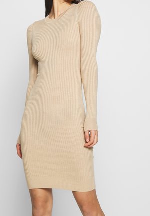 Mujer con vestido beige de punto acanalado, de manga larga y cuello redondo, de pie frente a un fondo claro y liso.
