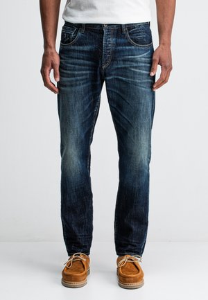 Denim jeans in dark blue wash, slim fit with faded texture, classic five-pocket design, and gold stitching. Paired with brown suede shoes.