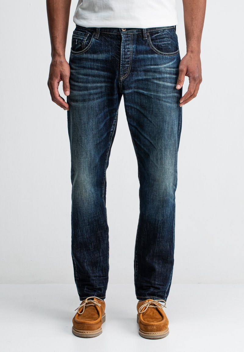 Denim jeans in een donkere blauwe wastint, slim fit met een vervaagde structuur, klassiek vijf-pocket ontwerp en gouden stiksels. Gepaarde met bruine suède schoenen.