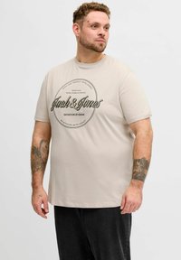 Lys beige bomuld T-shirt med korte ærmer. Har sort grafisk print med teksten "Jack & Jones" og en cirkulær ramme. Enkel design, afslappet pasform.