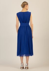 Blaues plissiertes Kleid mit taillierter Passform und ärmellosem Design. Mit einem Reißverschluss auf der Rückseite und einem halbtransparenten Saum. Silbergraue Absatzsandalen.