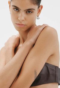 Top sin tirantes de cuero marrón con textura suave, brazos de la modelo cruzados, mostrando un tatuaje en el brazo interior, fondo neutro.