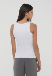 Biała bezrękawnik z gładkiego, elastycznego materiału, z okrągłym dekoltem, noszony z szarymi spodniami w talii, widok z tyłu.