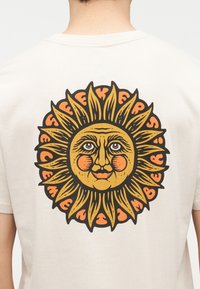 Πίσω μέρος λευκού πουκάμισου με μεγάλο εικονογραφημένο πρόσωπο ήλιου σε κίτρινο και πορτοκαλί, περιβαλλόμενο από κυκλικό, καμπυλωτό κείμενο.