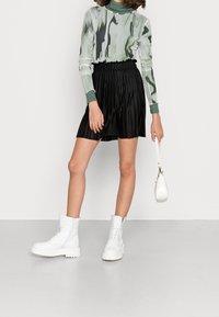 Haut à manches longues avec un imprimé abstrait vert et blanc, associé à une mini-jupe plissée noire. Porté avec des bottines blanches à lacets et un petit sac blanc.