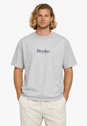 Mann mit schulterlangem blondem Haar, der ein graues T-Shirt mit dem Schriftzug "Broke. on Sundays" und helle gestreifte Hosen trägt, die Hände in den Taschen.