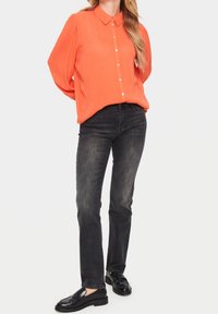 Frau mit einer leuchtend orangefarbenen Bluse mit Knöpfen, engen grauen Jeans und schwarzen Loafers, die mit den Händen hinter dem Rücken steht.