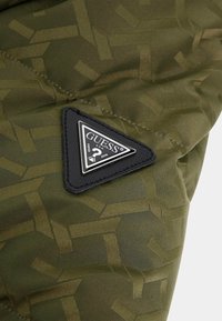 Green quilted fabric with an abstract pattern, featuring a black triangular logo patch labeled "GUESS." Texture appears smooth and durable.