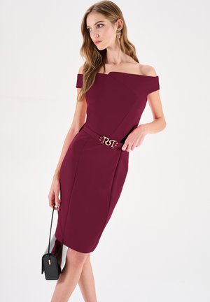 Robe bordeaux à épaules dénudées avec une silhouette ajustée, un design asymétrique et une ceinture accentuée dorée. Cheveux longs et ondulés ainsi qu'un sac à main noir complètent le look.