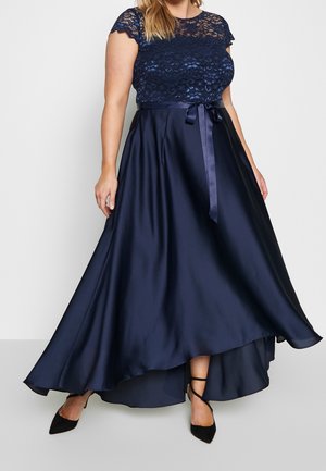 Plus-size vrouw draagt een marineblauwe kanten topjurk met een satijnen high-low rok, een striklint in de taille en zwarte hoge hakken met enkelriempje.