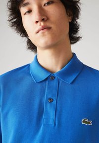 Niebieska koszulka polo z materiału bawełnianego o strukturalnej fakturze, z klasycznym kołnierzykiem, posiadająca dwa guziki oraz małe, haftowane logo krokodyla na piersi.
