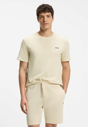 Mężczyzna w beżowej, teksturowanej koszuli z krótkim rękawem oraz pasujących szortach z troczkiem i logo "BOSS" na piersi i udzie.