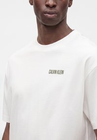 Άνδρας που φοράει λευκό T-shirt Calvin Klein με μικρό λογότυπο της μάρκας στο αριστερό στήθος, εμφανιζόμενος από το λαιμό μέχρι το μέσο του κορμού σε απλό φόντο.