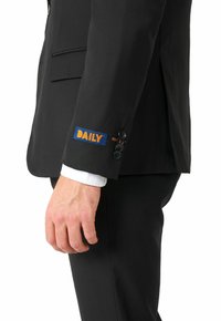 Chaqueta de traje negra con un parche azul en la manga, que presenta el texto "DAILY" en naranja. Incluye botones negros y detalles en los puños.