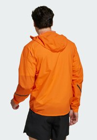 Veste légère orange avec capuche, présentant une texture lisse, des poignets élastiques, et des accents réfléchissants sur les manches. Vue arrière.