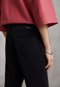 Černé manšestrové kalhoty s vertikálními žebry, s dvěma zadními kapsami a tkanou etiketou na pasu. Doplněné červenou košilí.