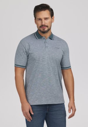 Mężczyzna w szarej koszulce polo z krótkim rękawem, z ciemnozielonym kołnierzem i mankietami, stojący na jednolitym jasnym tle.