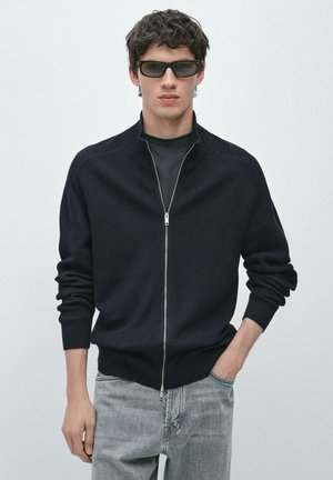 Giovane uomo con maglione nero con zip, t-shirt nera, jeans grigi e occhiali da sole, in piedi davanti a uno sfondo chiaro uniforme.