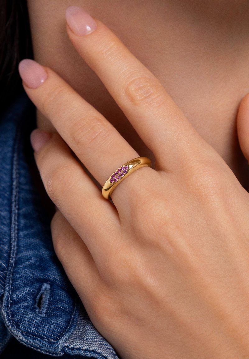Anillo de oro con una banda lisa que presenta una fila de piedras preciosas rosas. Mano mostrada con uñas naturales, usando una chaqueta de mezclilla.