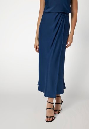 Femme portant une robe midi bleu marine avec un détail drapé, associée à des sandales noires à talons hauts et fines lanières, sur un fond neutre.