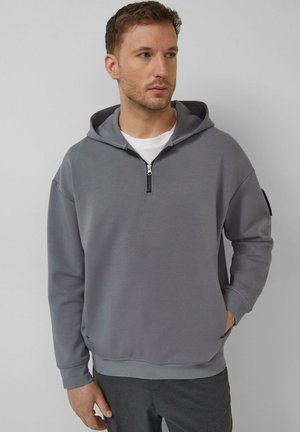 Mann trägt einen grauen Kapuzenpullover mit einem Halb-Reißverschluss, ein weißes Unterhemd und dunkelgraue Hose, steht vor einem einfarbigen Hintergrund.