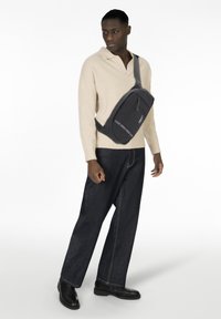 Uomo che indossa un maglione beige e jeans scuri, con una borsa a tracolla nera con logo visibile sul davanti, in piedi contro uno sfondo bianco.