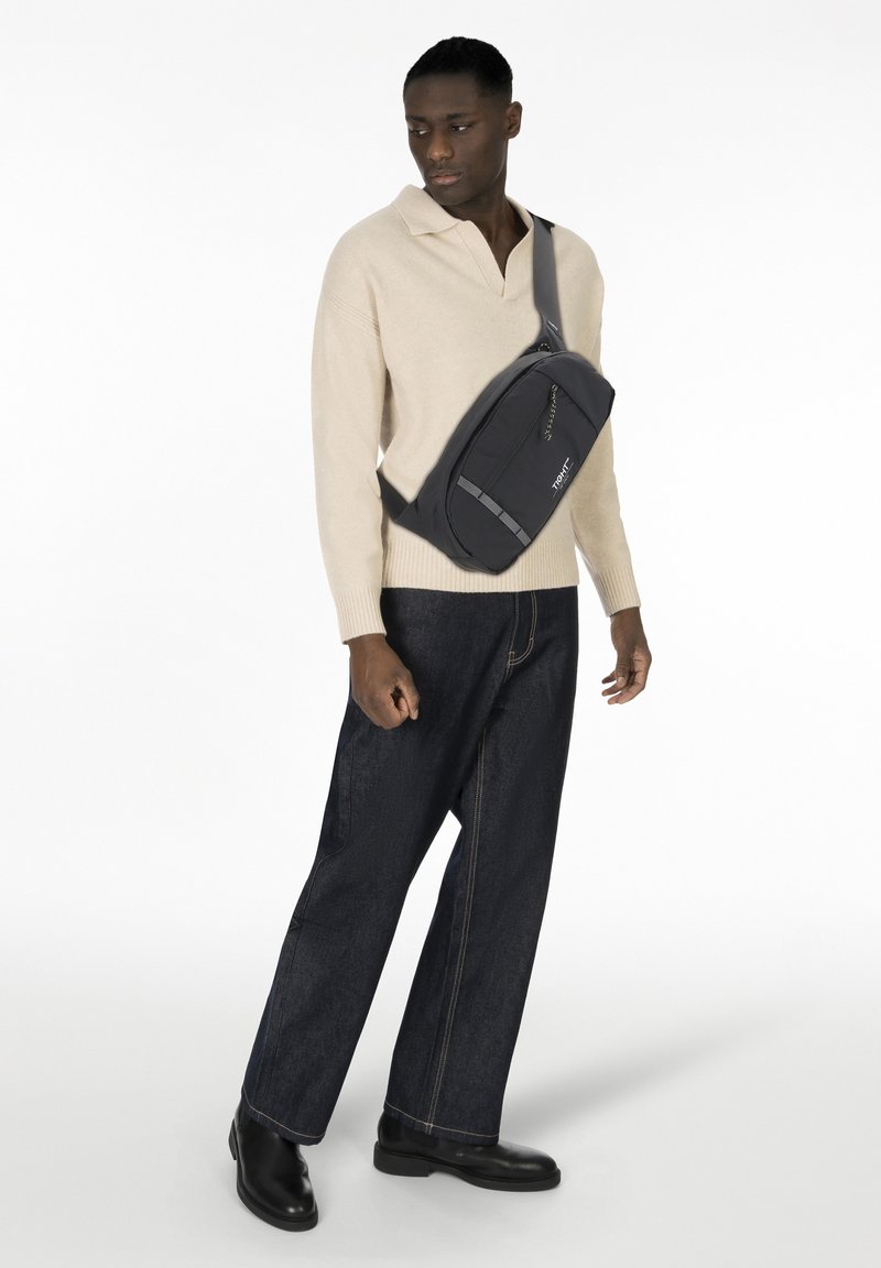 Uomo che indossa un maglione beige e jeans scuri, con una borsa a tracolla nera con logo visibile sul davanti, in piedi contro uno sfondo bianco.