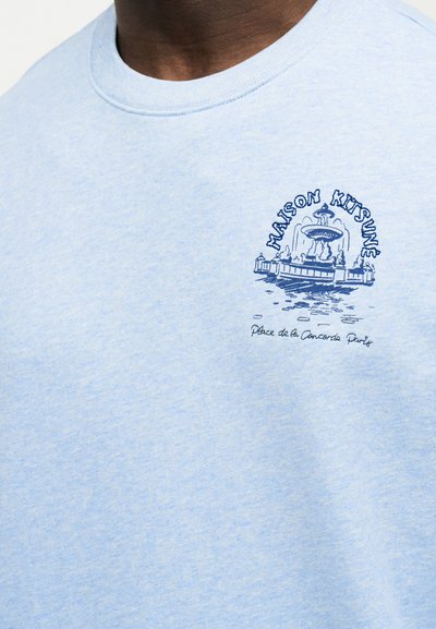 Φωτεινό μπλε πουκάμισο με μπλε γραφικό συντριβανιού και κείμενο "MAISON KITSUNE" και "Place de la Concorde Paris" στο στήθος.