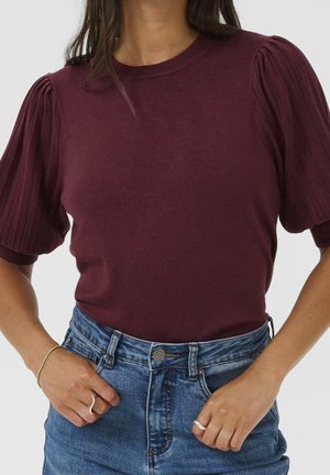 Femme portant un haut en maille bordeaux à manches courtes bouffantes, associé à un jean en denim bleu taille haute, accessoirisé avec des bracelets et des bagues.