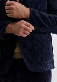 Handen die de knoop van de manchet van een donker marineblauwe corduroy blazer dichtknopen, gedragen over een tan kleurige trui.