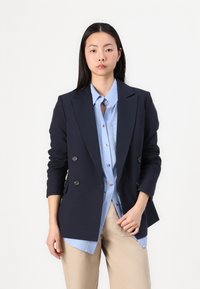 Blazer doppiopetto blu navy con due bottoni, rever a rotondo e forma aderente. Indossato sopra una camicia azzurro chiaro con bottoni e pantaloni beige.