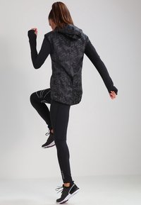 Czarna sportowa kamizelka z kapturem, ozdobiona wzorem kamuflażu, połączona z czarnymi legginsami i sportowymi butami. Skupienie na ruchu i projektowaniu odzieży aktywnej.