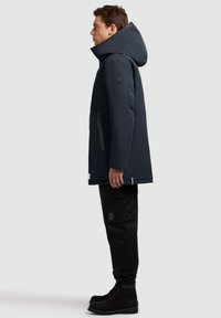 Mørkeblå isoleret jakke med stor hætte, sidelomme med lynlås og tekstureret stof. Suppleret med sorte cargo bukser og sorte ruskindsstøvler.